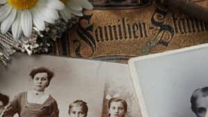 A imagem mostra uma série de objetos antigos dispostos sobre uma mesa. Um livro de família ao centro fotos familiares em preto e branco abaixo e uma flor de pétalas brancas a esquerda.