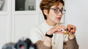 A imagem mostra uma mulher de cabelos curtos e ondulados. Ela usa óculos de grau e uma blusa branca de gola alta. Ela está em um ambiente que se parece a um escritório. Está sentada, com os cotovelos apoiados em uma mesa e com a boca semiaberta como se estivesse conversando com alguém.