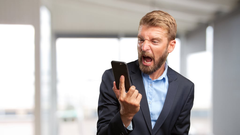 Um homem branco, loiro e bem vestido com um terno azul marinho com camisa de botões azul claro olhando e gritando irado enquanto olha para um celular que seguram com a mão direita.