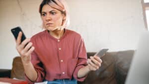 A imagem mostra uma mulher com uma expressão de preocupação. Ela tem cabelo curto loiro, usa um piercing no nariz e está vestindo uma camisa de botões rosa. A pessoa está segurando um smartphone em cada mão e parece estar muito preocupada com algum problema inesperado nos dispositivos.