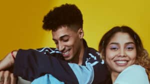 Um casal jovem e sorridente posa juntos contra um fundo amarelo vibrante. O homem, pardo, com cabelo encaracolado curto e uma camiseta escura, descansa sua cabeça levemente sobre o ombro da mulher. A mulher, exibindo cachos soltos e uma blusa de cor clara, Ambos exalam uma sensação de conforto e afeto compartilhado, sugerindo uma conexão próxima e um espírito jovial.