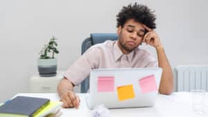 A imagem mostra um homem de pelos crespos e pretos e camisa bege. Ele apoia a cabeça em uma das mãos e exprime um ar de preocupação olhando para a tela de um computador. Ele está sentando em uma cadeira em um ambiente que parece um escritório. A sua frente estão algumas pastas e cadernos.