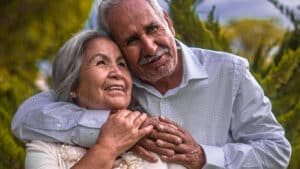 A imagem mostra duas pessoas idosas, um homem e uma mulher, ambos de cabelos brancos. O homem passa o braço pelo pescoço da mulher e entrelaça os dedos no seu ombro. A mulher apoia a sua mão na mão do homem. Os dois olham para a frente e sorriem.