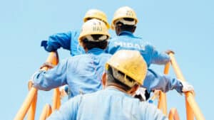 A imagem mostra quatro trabalhadores. Todos usam macacão azul de mangas compridas e capacete amarelo. Eles estão em fila, subindo por uma escada de corrimão laranja.