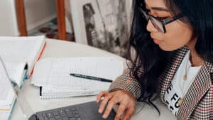 A imagem mostra uma mulher de cabelos longos e escuros. Ela usa óculos de grau e olha para a tela de um computador. Com a mão direita, digita algo no teclado. Ao lado do computador estão cadernos, lápis e canetas, todos apoiados em uma mesa.