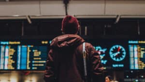 A imagem mostra um homem de costas. Ele veste uma blusa de frio com capuz de cor vinho e um gorro da mesma cor. Pendurado no seu ombro direito, uma câmera fotográfica. Ao fundo, um grande painel com leyras e números iluminados indicando voos.