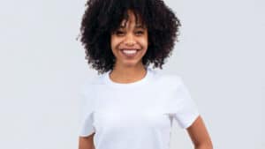 A imagem mostra uma mulher de cabelos crespos e soltos, sorrindo para a imagem. Ela usa uma blusa de manga curta branca e está em um ambiente com fundo branco.