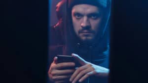 A imagem mostra um homem com moletom e capuz, olhando fixamente para a câmera e com seriedade. Com as mãos, ele mexe com um celular, como se cometesse um golpe de estelionato. Ele está em um ambiente escuro e sombrio, e vemos o seu rosto em uma fresta entre dois computadores.