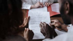A imagem mostra quatro mãos segurando um documento que contém algumas assinaturas. O documento está próximo de uma mesa, do outro lado da mesa aparece parte do corpo de uma mulher que parece esperar pela leitura do casal que mantém as mãos no documento.