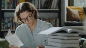 A imagem mostra um homem de cabelos medianos e loiros com óculos de grau e camisa social cinza. Ele está sentado em um local qye se parece a uma biblioteca e, a sua frente, estão vários livros. Uma das mãos segura um lilvro aberto e a outra seguro um lápis, que ele usa para escrever em um papel.