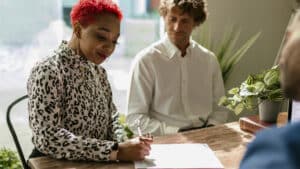 A imagem mostra uma mulher negra de blusa de manga comprida e cabelos vermelhos. Ela está sentada e assina um documento apoiado em uma mesa de madeira. Ao fundo, um homem de blusa social acompanha a ação. De frente para eles, um homem de blazer azul, do outro lado da mesa, aguarda a assinatura.