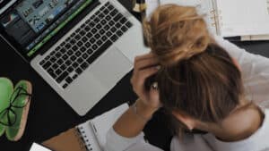 A imagem mostra uma mulher vista de cima. Conseguimos ver que ela está sentada e em frente a um computador. Ao seu lado tem um óculos de grau e um caderno de anotações aberto. Ela está com os cabelos presos em um coque e as mãos na testa, como se algo estivesse dando errado.