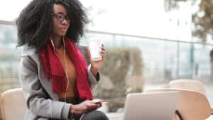 A imagem mostra uma mulher de pele negra, cabelos longos, crespos e soltos, usando um óculos de grau e roupas de frio. Ela está em um ambiente externo. Com a mão esquerda segura um copo de café e, com a mão direita, um celular, com o fone conectado aos ouvidos. Ela olha para um computador que está apoiado em uma mesa a sua frente.