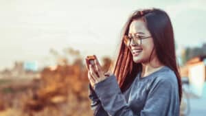 A imagem mostra uma mulher de cabelos lisos e soltos e óculos de grau. Ela está de lado e segura o celular com as duas mãos. Ela olha para o celular e sorri.