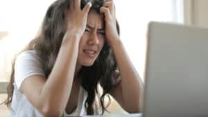 A imagem mostra uma mulher de cabelos longos sentada e olhando para um computador. Ela está com as mãos na testa e com uma expressão de sofrimento.