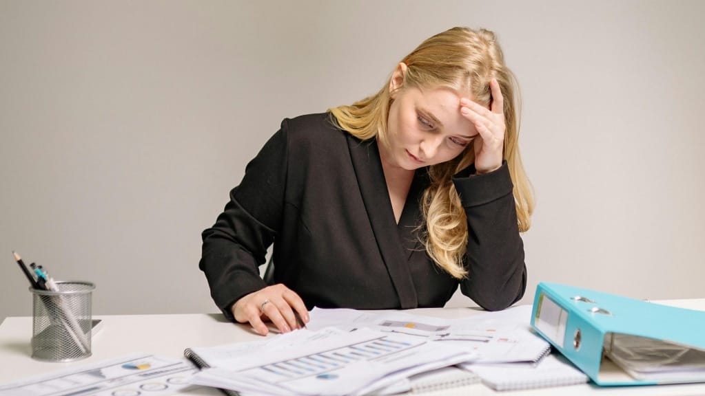 A imagem mostra uma mulher de pele branco e cabelos loiros, longos e soltos. Ela está sentada em uma cadeia, em frente a uma mesa com diversos cadernos e papéis a frente. Sua mão esquerda está apoiando a testa e ela parece preocupada.