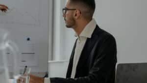 A imagem mostra um homem de cabelo raspado, blusa social, casaco e óculos de grau. Ele está em um escritório, sentado em uma cadeira com uma mesa a frente. Ele olha para a direita, como se estivesse conversando com alguém.