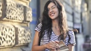A imagem mostra uma jovem de cabelos pretos, ondulados e soltos. Ela usa óculos de grau e uma blusa de manga curta listrada de preto e branco. Ela segura livros com as duas mãos e sorri para a foto. Ela está ao lado de um muro de pedra, em um espaço aberto.