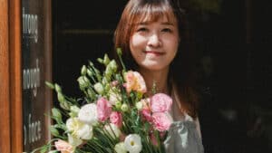 A imagem mostra uma mulher de cabelos marrons e lisos, com franja. Ela carrega, a frente do seu corpo, um buquê de flores coloridas. Ela está posicionada em frente a uma porta aberta e sorri para a câmera.