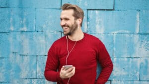 A imagem mostra um homem de pele branca, cabelos loiros e curtos e barba feita. Ele usa uma blusa vermelha de manga comprida e fones de oouvidos que se conectam a um celular que está nas suas mãos. Ele está apoiado em um muro azul e sorri com o rosto levemente virado para a direita.
