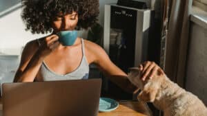 A imagem mostra uma mulher negra de cabelos crespos e soltos. Ela está sentada em uma mesa em uma área com sol. Ela está com o computador aberto a sua frente. Com uma das mãos, bebe algo em uma xícara de café de cor azul e, com a outra mão, faz carinho em um cachorro.