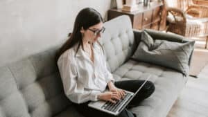 A imagem mostra uma mulher de cabelos lisos e semi presos e óculos de grau. Ela veste uma calça preta e uma blusa de manga comprida branca; Está sentada em um sofá cinza, com as pernas cruzadas e um computador apoiado nas pernas. Ela digita neste computador com as duas mãos enquanto olha para a tela.