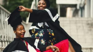A imagem mostra duas mulhere negras em uma festa de graduação. Elas estão em um espaço aberto e usam uma beca, roupa preta típica das pessoas que estão se formando. Uma delas está sentada e a outra brinca com ela colocando o chapéu da beca no alto da sua cabeça.