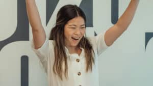 A imagem mostra uma jovem de pele clara. Seus cabelos longos e semi-preso caem em cima dos ombros. Ela usa uma blusa branca com botões marrons. Ela está sorrindo, com os olhos fechados e os braços para cima, como se estivesse vibrando.