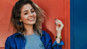 A imagem mostra uma jovem de cabelos loiros, ondulados e soltos, usando uma camiseta cinza e um casaco jeans. Ela está em fente a uma porta laranja. Ela sorri para a câmera enquanto mexe na ponta dos cabelos com a mão esquerda.