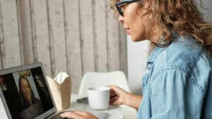 A imagem mostra uma mulher de pele branca, cabelos enrolados, loiros e soltos, usando óculos de grau e uma camisa jeans de manga comprida. Vemos esta mulher de perfil. Ela está sentada em frente a um computador, com a tela aberta, onde é possível ver outra pessoa em uma chamada de vídeo. A mulher olha para a tela, segura uma caneca com a mão direita e tecla com a mão esquerda.