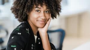 A imagem mostra uma mulher de pele negra, com cabelos crespos e soltos, em estilo black power. Ela usa uma blusa branca por baixo e uma blusa preta com deatalhes brancos por cima. Sua mão esquerda apoia o queixo enquanto ela sorri e olha para a câmera.