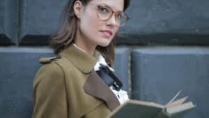 A imagem mostra uma mulher branca de cabelos lisos até a altura dos ombros e solto. Ela usa óculos de grau e veste uma blusa branca e um casaco verde escuro. Ela está com um livro aberto e olha para a câmera.