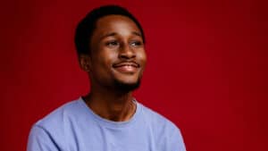A imagem mostra um jovem negro de cabelo pretos e curtos. Ele usa uma camiseta azul e olha para o lado direito com um sorriso no rosto e um olhar confiante. Ele está em uma sala com uma parede vermelha.