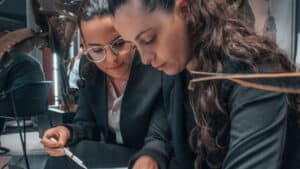 A imagem mostra duas mulheres brancas. Ambas têm cabelos longos, marrons e presos, usam blusa branca e terno preto. Elas estão sentadas lado a lado, com a cabeça baixa enquanto leem um documento.