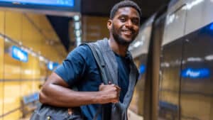 A imagem mostra um jovem negro de cabelos pretos e curtos. Ele usa uma blusa azul escura e carrega uma mochila nas costas, enquanto segura uma das alças com as mãos. Ele conseguiu um auxílio-deslocamento e está em um corredor de aeroporto. Ele sorri para a foto.