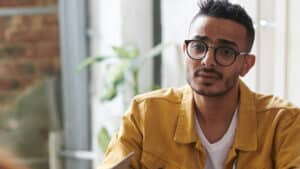 A imagem mostra um homem de cabelos pretos e curtos usando um óculos de grau, camisa branca e casaco amarelo. Ele está sentado dentro de um escritório e olha para um interlocutor com uma expressão de incompreensão.
