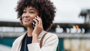 A imagem mostra uma mulher negra de cabelos crespos, curtos e soltos. Ela usa uma blusa preta e um casaco bege por cima. Ela está em um espaço ao ar livre, uma das mãos segura um telefone no seu ouvido. Ela olha para a frente com um sorriso.