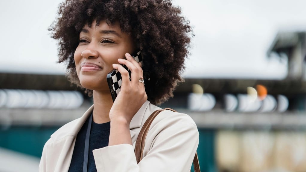 A imagem mostra uma mulher negra de cabelos crespos, curtos e soltos. Ela usa uma blusa preta e um casaco bege por cima. Ela está em um espaço ao ar livre, uma das mãos segura um telefone no seu ouvido. Ela olha para a frente com um sorriso.