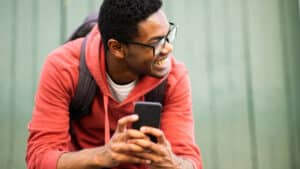 A imagem mostra um jovem negro de cabelos pretos e curtos, usando um óculos de grau, um moletom vermelho e uma mochila nas costas. Ele está apoiado em um aparador, com um celular nas mãos. Ele olha para o lado direito e sorri.