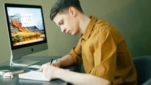 A imagem mostra um jovem branco de cabelo curto. usando uma camisa de gola de cor mostrada. Ele está sentado em uma cadeira e escreve em um caderno com apoiado em uma mesa. A sua frente tem um computador e materiais de estudo.