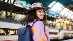 A imagem mostra uma mulher jovem de cabelos enrolados, medianos e soltos. Ela usa um chapéu de palha, uma blusa de frio listrada de laranja e lilás e uma bolsa preta apoiada nas costas. Ela está de costas, mas seu rosto está voltado para a câmera e ela sorri. Ela está em uma estação de trem.