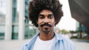 A imagem mostra um homem jovem negro, de cabelo black power e bigode. Ele usa uma blusa branca e uma camisa social azul clara por cima. O homem está em um espaço aberto e olha para a câmera.
