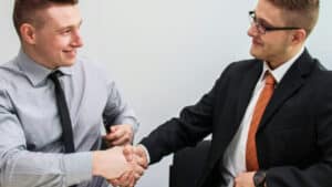 A imagem mostra dois homens loiros de cabelo curto olhando um para o outro enquanto apertam as mãos definindo o tipo societário da empresa. O homem da esquerda usa uma blusa social cinza e uma gravata vinho e o homem da direita usa uma blusa social branca, um paletó preto e uma gravata laranja.