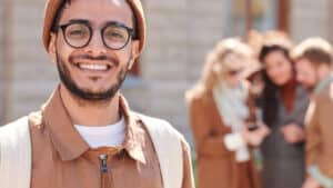 A imagem mostra um homem jovem, branco de barba e bigode, usando óculos de grau e uma touca escondendo os cabelos. Ele usa uma blusa branca e uma camisa social marrom por cima. Está olhando para a cÊmera e sorrindo, ao lado esquerdo da imagem. À direita, ao fundo, um grupo de três pessoas olham para uma imagem em um celular, compondo o quadro. Todos estão em um espaço aberto e iluminado pelo sol.