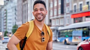 A imagem mostra um homem de pele clara, cabelos curtos e pretos. Ele usa uma blusa laranja, uma mochila de alças amarelas e um óculos preto pendurado na blusa. Ele sorri enquanto olha para a esquerda sorrindo. Ele está em uma rua, com veículos passando e prédios ao fundo.