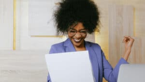 A imagem mostra uma mulher negra de cabelos crespos e soltos, usando um paletó azul claro. Ela está em uma sala, sentada em uma cadeira a frennte de uma mesa. Ela olha para um documento enquanto sorri.