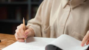 A imagem mostra uma mulher sentada em uma cadeira com um caderno de provas a frente com a mão em uma caneta apoiada neste caderno, como se estivesse fazendo uma correção. Ela usa uma blusa de manga comprida bege.