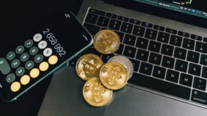 A imagem mostra uma mesa onde vemos um computador aberto, um celular no aplicativo de calculadora e moedas prateadas e douradas representando criptomoedas.