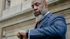 A imagem mostra um homem negro, careca, com barba grisalha. Ele usa um paletó azul marinho e uma gravata de cor vinho. Ele está em um espaço aberto e olha para a hora no seu relógio de pulso.
