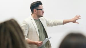A imagem mostra um professor em sala de aula. Ele está a frente da turma, virado para a direita com o braço esquerdo levantado e o direito segurando um caderno. Ele é um homem branco, de cabelos curtos, usando óculos de grau, blusa verde escura e paletó branco.